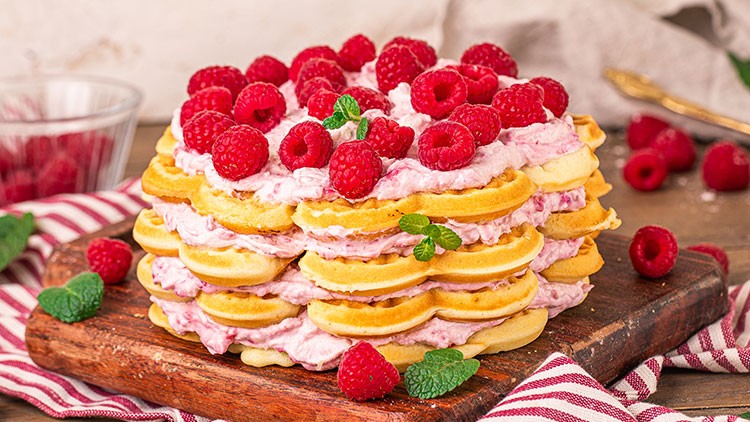 Ein gestapelter Waffelkuchen mit Schichten aus Himbeercreme, frischen Himbeeren und Minzblättern auf einem Holzbrett.
