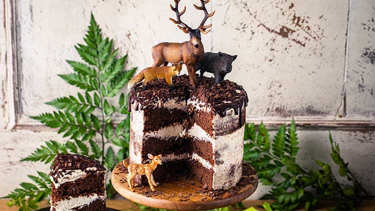 Schichttorte mit Schokoladenstücken, Schokoglasur und Miniatur-Waldtieren aus Plastik auf einem Holzteller, umgeben von Grünpflanzen.