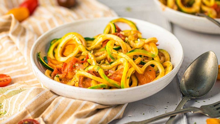 Ein weißer Teller mit Zucchininudeln, Tomatenstücken und Gewürzen auf einem gestreiften Tuch, daneben liegen Löffel und Gabel.
