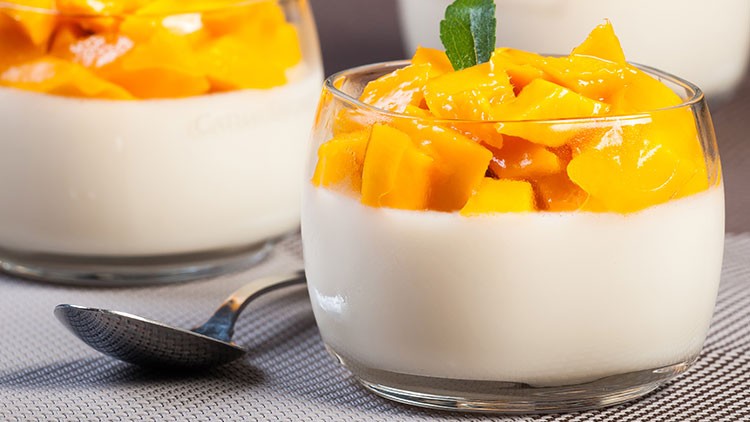 Ein Glas mit weißem Pudding, darauf frische Mangostücke und ein grünes Minzblatt, daneben ein silberner Löffel.