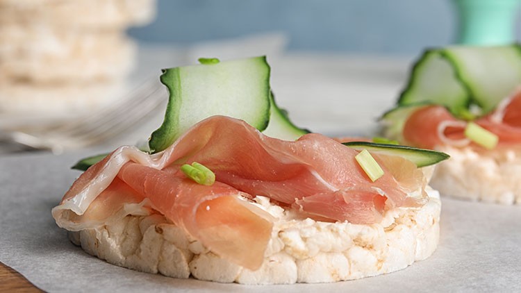 Reiswaffel belegt mit Schinken, Gurkenscheiben und kleinen grünen Zwiebelfrühlingsstücken auf hellem Untergrund.