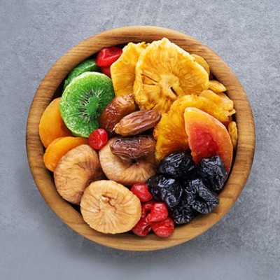 Runde Holzschale mit getrocknetem Obst wie Ananas, Feigen, Pflaumen, Aprikosen, Kiwi, Datteln und roten Beeren auf grauem Untergrund.