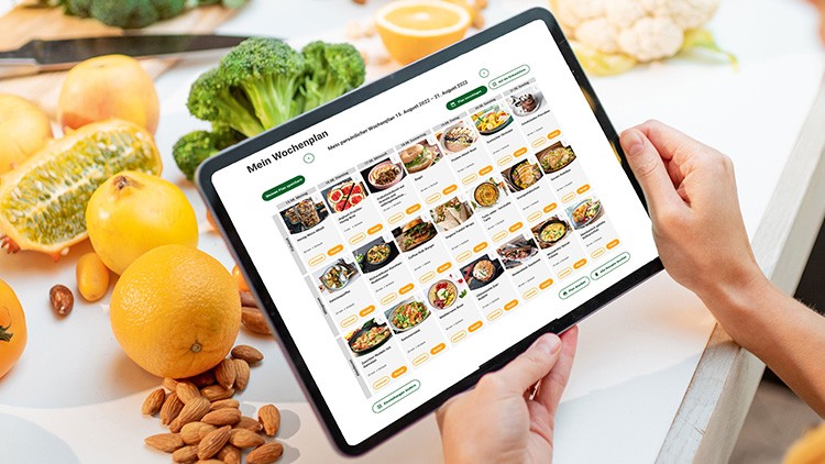 Eine Person hält ein Tablet mit der geöffneten Ansicht „Mein Wochenplan“, im Hintergrund liegen Gemüse, Obst und Nüsse.