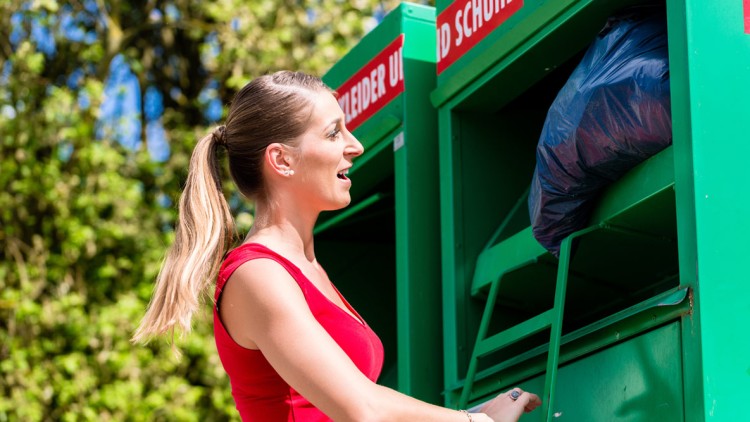 Person in rotem Oberteil legt eine gefüllte blaue Plastiktüte in einen grünen Container für Kleidung und Schuhe.