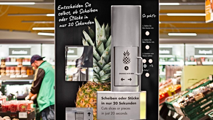 Ananas-Schneidemaschine mit Anleitung und Schriftzug „Scheiben oder Stücke in nur 20 Sekunden“, im Hintergrund ein Supermarkt.
