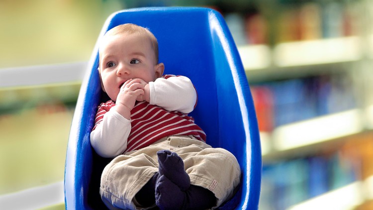 Kind in beige Hose, dunklen Socken und rot-weiß gestreiftem Oberteil sitzt in einem blauen Einkaufswagensitz.
