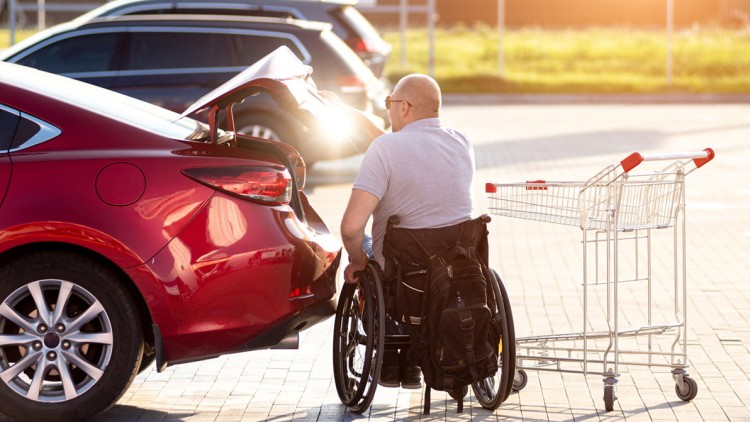 Person im Rollstuhl vor offenem Kofferraum eines roten Autos, daneben ein leerer Einkaufswagen, Sonnenlicht im Hintergrund.