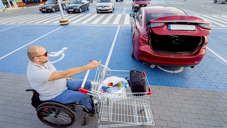 Ein Mann im Rollstuhl mit Einkaufswagen ist auf einem Parkplatz mit Rollstuhlsymbol neben einem roten Auto mit offenem Kofferraum.