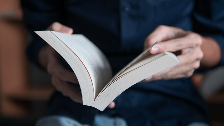 Person in dunkler Kleidung hält ein aufgeschlagenes Buch, während die Seiten durchgeblättert werden.