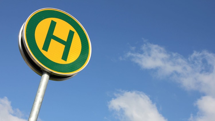 Rundes gelb-grünes Haltestellenschild mit einem grünen „H“ vor blauem Himmel mit wenigen weißen Wolken.