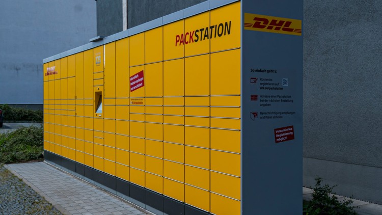 Gelbe DHL-Packstation mit mehreren Fächern und grauer Infotafel, darauf Anweisungen zur Nutzung und ein rotes Hinweisfeld.