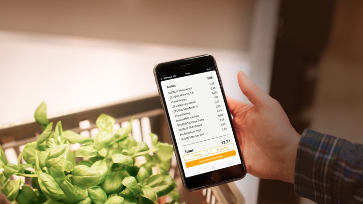 Eine Hand hält ein Smartphone mit geöffneter mein GLOBUS App, ein Einkaufswagen mit frischem Basilikum steht im Vordergrund.