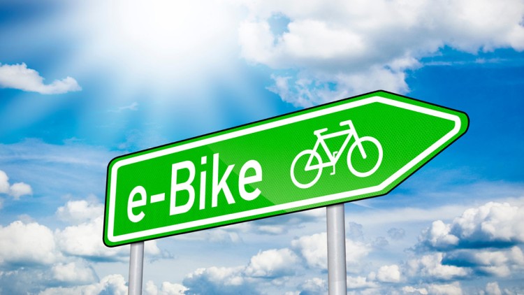 Grünes Schild mit der Aufschrift e-Bike, einem Fahrradsymbol und blauem Himmel mit Sonne und Wolken im Hintergrund.