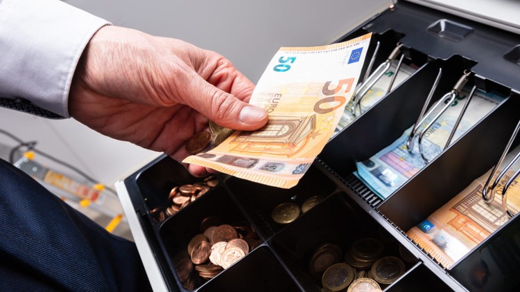 Eine Hand legt einen 50-Euro-Schein in eine geöffnete Kassenlade mit mehreren Fächern für Münzen und Banknoten.