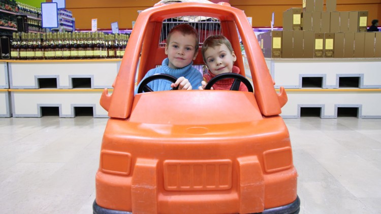 Zwei Kinder sitzen in einem orangefarbenen Spielauto mit schwarzen Lenkrädern in einem Laden vor Regalen und Kartons.