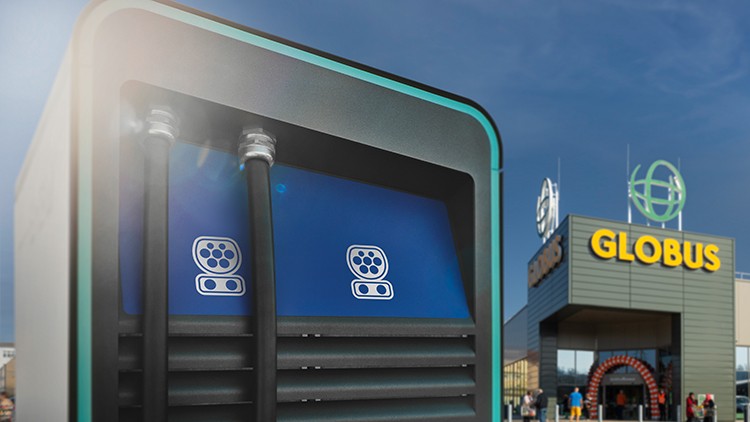 Ladestation mit zwei Kabeln und Steckersymbolen, im Hintergrund ein GLOBUS-Markt mit Logo und Eingang.
