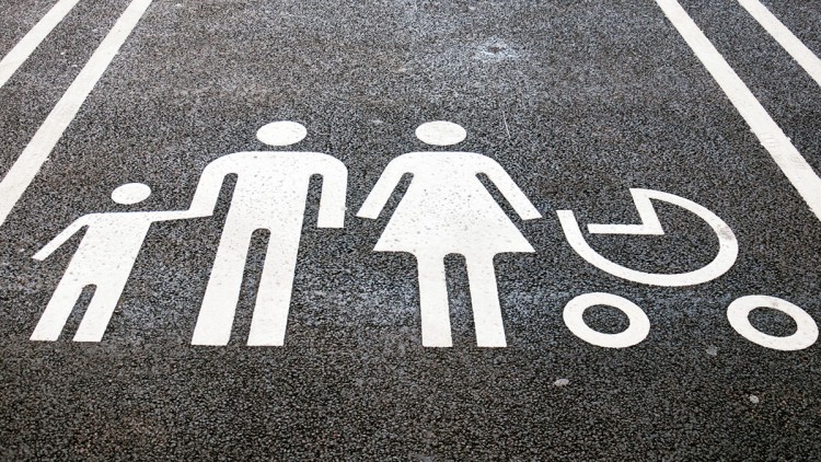 Weiße Bodenmarkierung auf Asphalt zeigt eine Gruppe aus drei Personen und einem Kinderwagen, flankiert von Parkstreifen.
