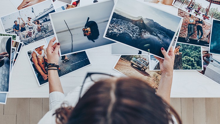 Person hält mehrere bunte Fotos in den Händen, die auf einem weißen Tisch verstreut und teilweise überlappend liegen.
