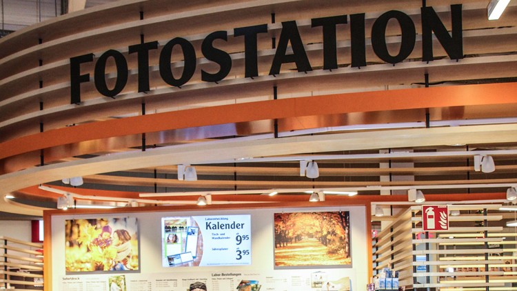 Fotoentwicklungsstation mit Schriftzug „FOTOSTATION“, Bildern, Texten zu Fotoprodukten und Bildschirmen in einem hellen Raum.