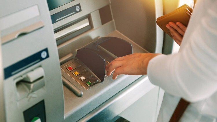 Eine Person bedient einen Geldautomaten, hebt die Hand zur Tastatur und hält eine braune Brieftasche in der anderen Hand.