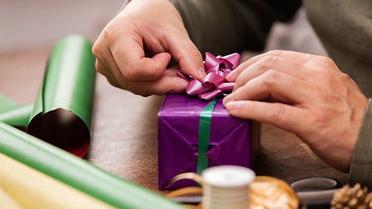 Hände binden eine pinke Schleife auf ein lila Geschenk mit grünem Band, daneben Geschenkpapier und Bastelutensilien.