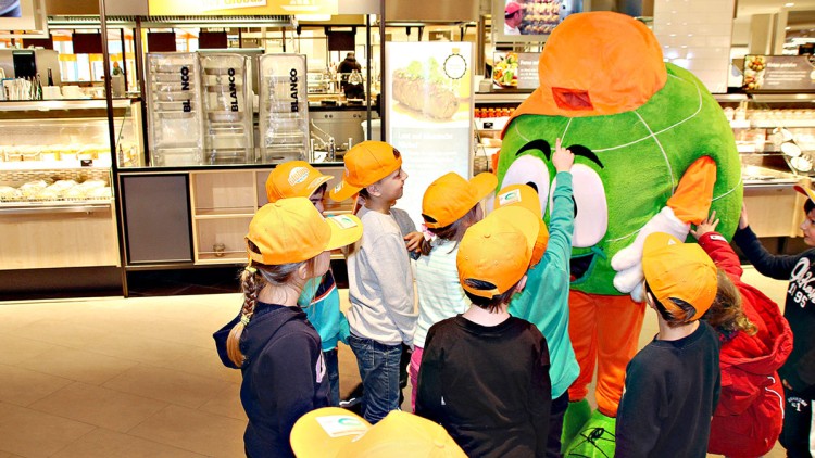 Kindergruppe mit orangefarbenen GLOBUS-Kappen steht vor Globini im Eingangsbereich eines GLOBUS-Markts.