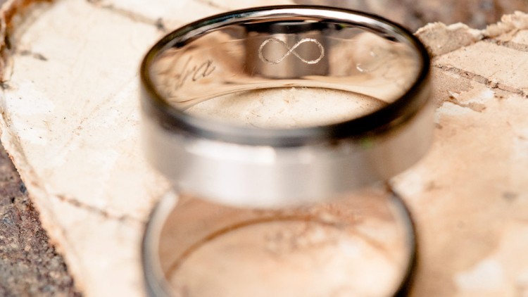 Zwei silberne Ringe mit schwarzem Rand auf hellem Untergrund, einer zeigt eine Gravur mit dem Symbol der Unendlichkeit.