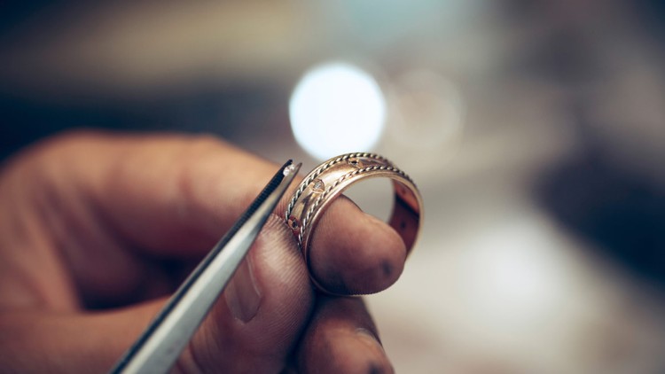 Eine Hand hält einen goldenen Ring mit eingraviertem Muster, während eine Pinzette einen kleinen Stein darüber positioniert.