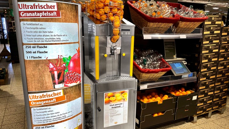 Saftpresse mit Orangen und Flaschen zur Selbstabfüllung, daneben Glaskaraffen und Hinweisschilder zu Granatapfel- und Orangensaft.
