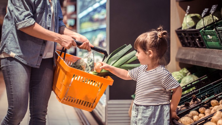 Eine Person mit oranger Einkaufskorb reicht einem Kind ein Gemüse im Obst- und Gemüsebereich eines Supermarkts.