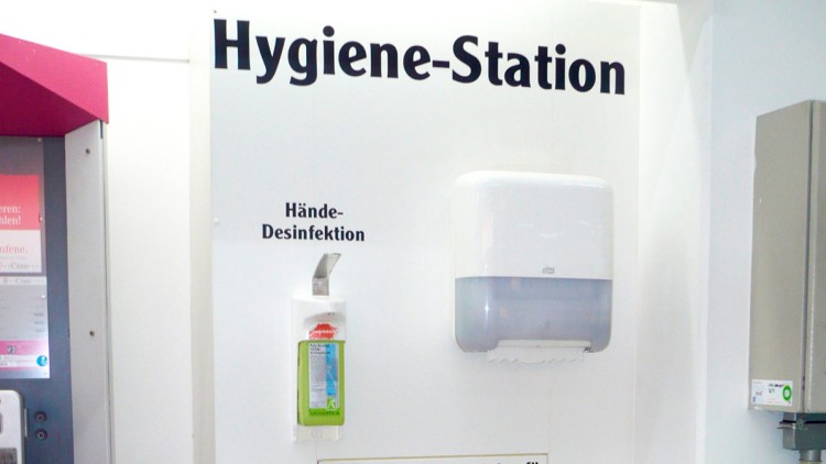 Eine weiße Hygiene-Station mit Spendern für Hände-Desinfektion, Einkaufswagendesinfektion und einer Papierhandtuchhalterung.