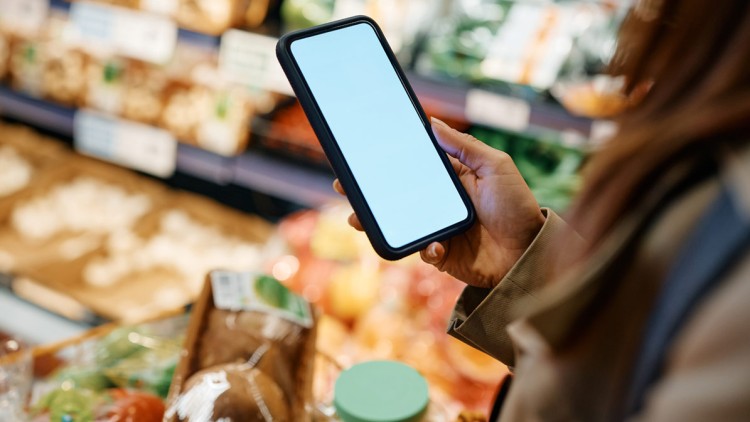Eine Hand hält ein Smartphone mit weißem Bildschirm über einem Einkaufswagen, im Hintergrund sind Obst und Gemüse.