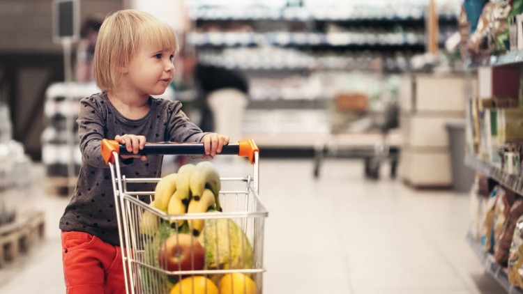 Kind schiebt einen Einkaufswagen mit Bananen, Melone, Äpfeln und weiteren Lebensmitteln durch einen Supermarktgang.