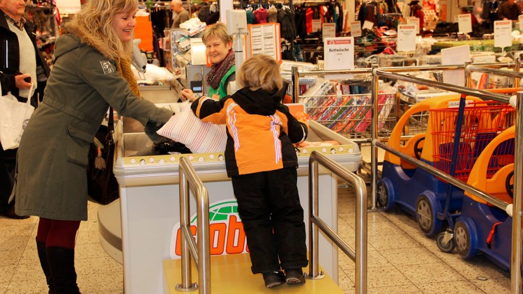 Ein Kind in orange-schwarzer Jacke steht an einem GLOBUS-Kassenband, daneben Einkäufe und Einkaufswagen im Hintergrund.