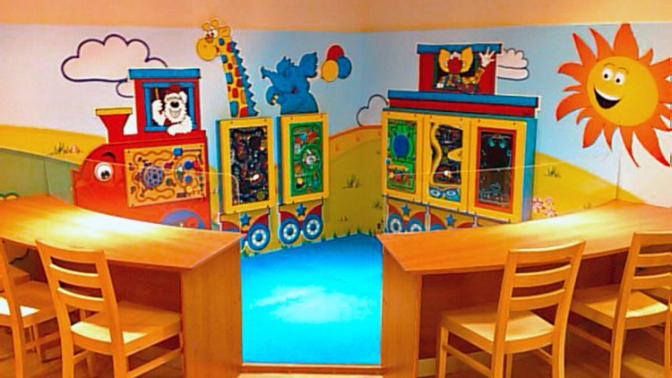 Bunter Spielbereich mit Holztischen und Stühlen vor einer bunten Wand mit einer bemalten Lokomotive, Tieren und Sonne.