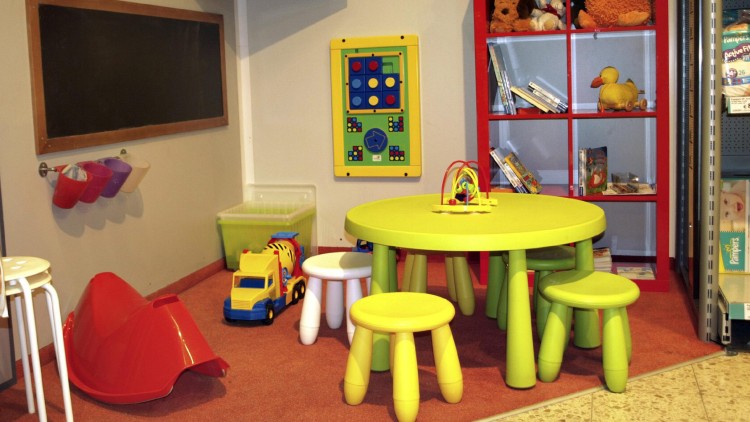 Spielecke mit grünem Tisch, gelben und weißen Hockern, Spielzeug wie LKW und Motorikschleife sowie Regal mit Büchern und Stofftieren.
