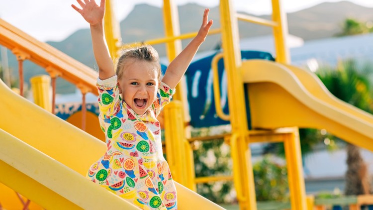 Kind rutscht mit erhobenen Armen eine gelbe Rutsche herunter, trägt ein buntes Kleid mit Obstmotiven, Spielplatz im Hintergrund.