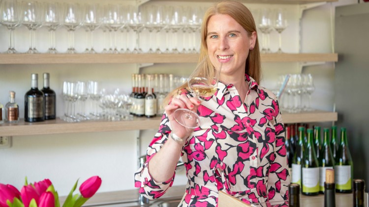 Person in gemustertem Kleid hält ein Glas Weißwein vor einer Regalwand mit Gläsern und Weinflaschen, vorn stehen Tulpen.