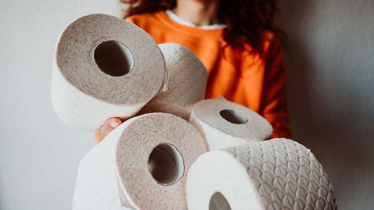 Person in orangem Oberteil hält mehrere Toilettenpapierrollen mit geprägtem Muster in den Händen.