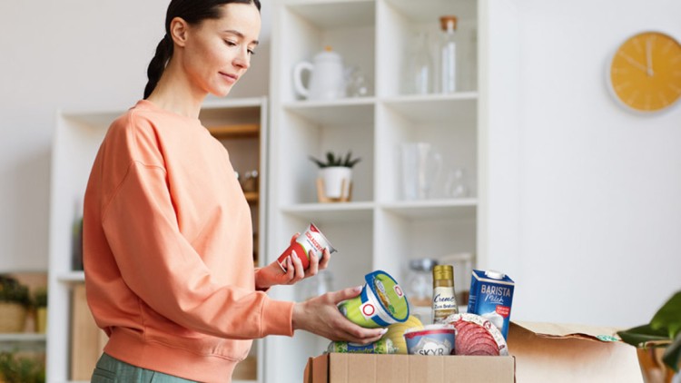 Person hält Lebensmittel in der Hand, daneben eine geöffnete GLOBUS Last-Minute-Box mit verschiedenen Produkten auf einem Tisch.