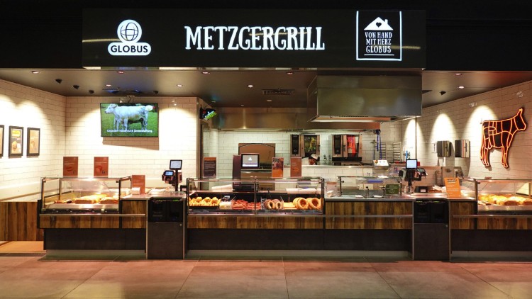 GLOBUS Metzgergrill mit beleuchteten Theken, Fleisch- und Wurstauslage, Werbetafel, Kuhbildschirm und orangener Neon-Kuhkontur.