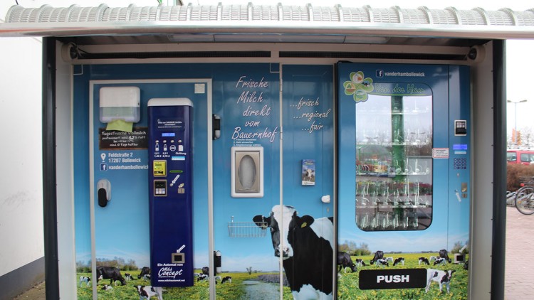 Ein Milchautomat mit blauer Front und Kuhmotiven, Werbung für „Frische Milch direkt vom Bauernhof“ und einer großen „PUSH“-Taste.