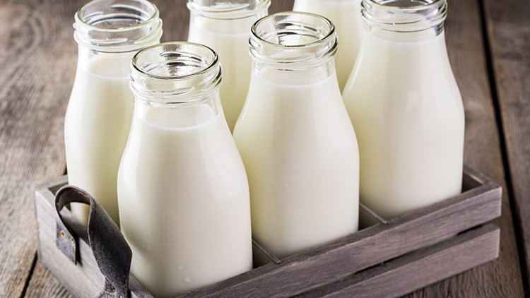 Sechs durchsichtige Glasflaschen mit Milch stehen in einer grauen Holzkiste auf einem Holztisch.