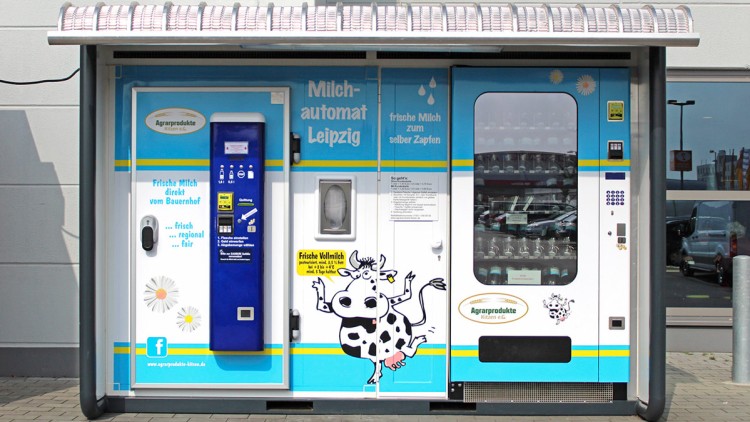 Blauer und weißer Milchautomat mit Aufschrift „Milchautomat Leipzig“ und Zeichnung einer Kuh, daneben Glasflaschenregal.