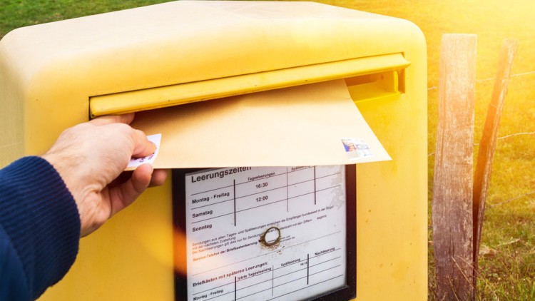 Eine Hand steckt einen Briefumschlag in einen gelben Briefkasten mit sichtbar angeschlagenen Leerungszeiten.
