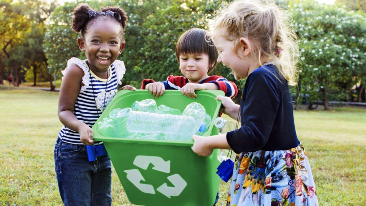 Drei Kinder tragen gemeinsam eine grüne Recyclingbox mit leeren Plastikflaschen auf einer Wiese im Freien.