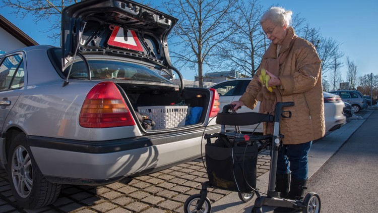 Silberfarbenes Auto mit offenem Kofferraum, darin Einkaufskorb, Person mit Rollator hält eine Banane.