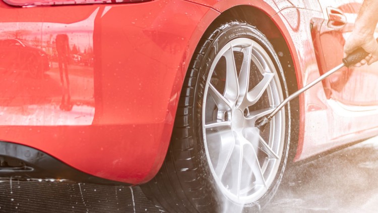 Eine Hand hält einen Hochdruckreiniger und säubert den silbernen Reifen eines roten Autos, während Wasser spritzt.