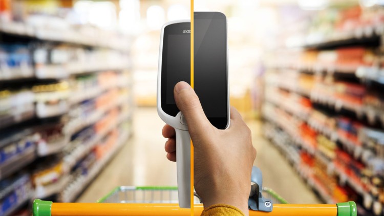Hand hält Scan & Go-Gerät über einem Einkaufswagen mit oranger Griffstange, im Hintergrund unscharfe Regale in einem Supermarkt.