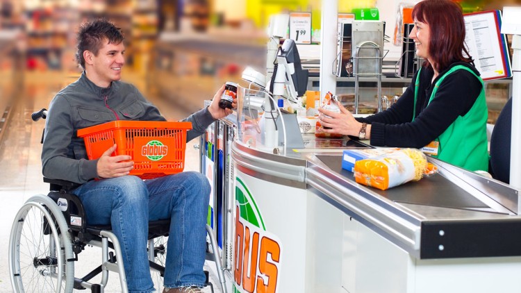 Mensch im Rollstuhl mit GLOBUS-Einkaufskorb reicht an einer Kasse ein Produkt an eine kassierende Person weiter.